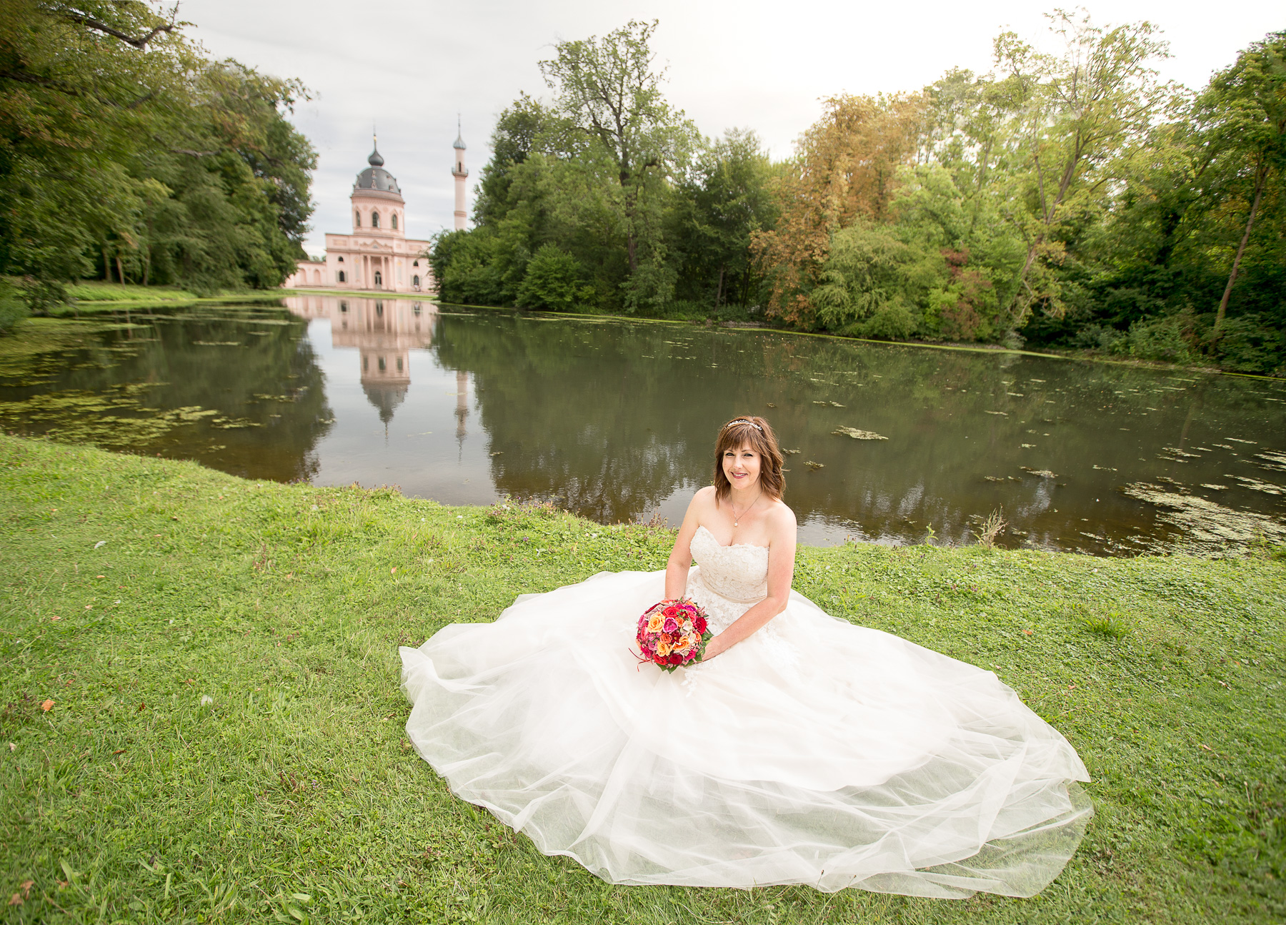Die Braut sitzt auf der Wiese des Schwetzinger Schlossgartens. Sie hat ein prächtiges Brautkleid an und hält einen Brautstrauß in ihren Händen. Im Hintergrund ist ein See und die Moschee des Schlossgartens sichtbar.
