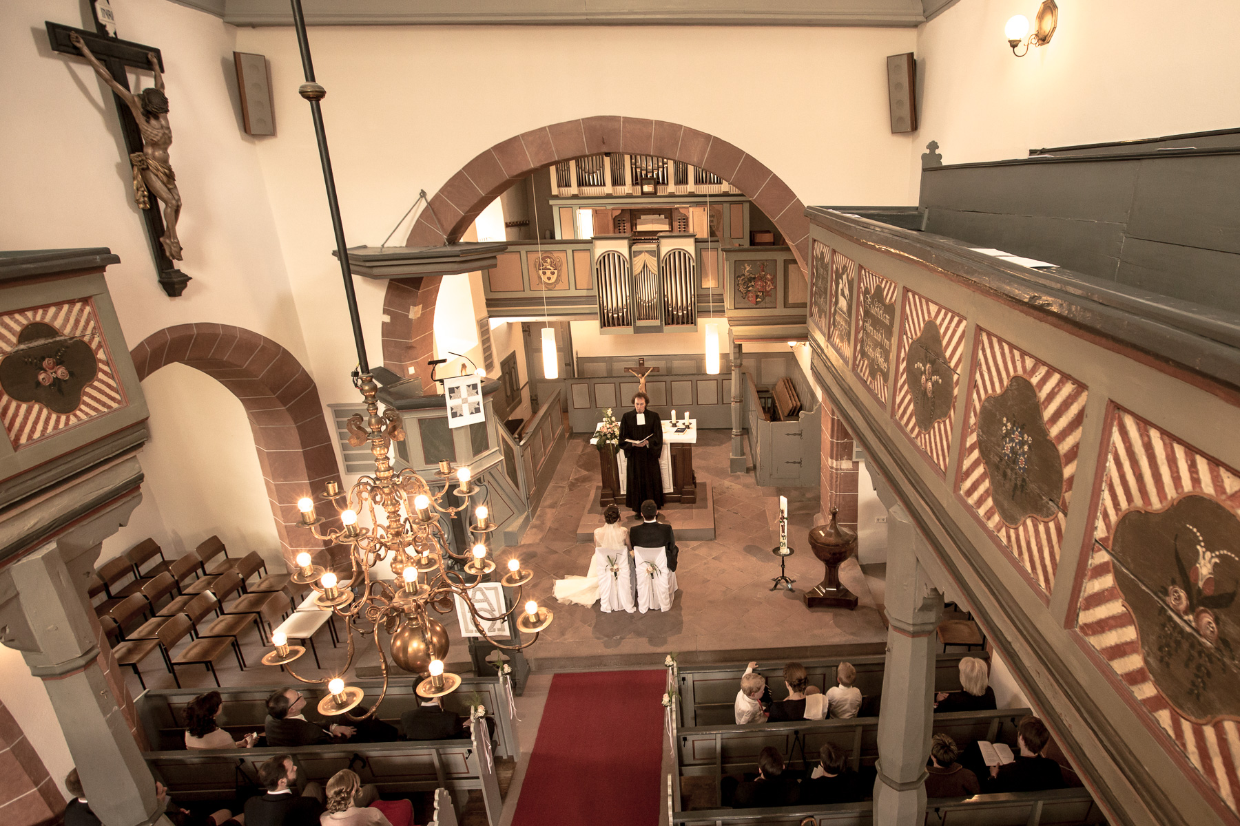 Ein Überblick des Kirchensaals von oben. Das Hochzeitspaar, der Priester vor ihnen und ein Teil der Gäste hinter ihnen sind erkennbar.