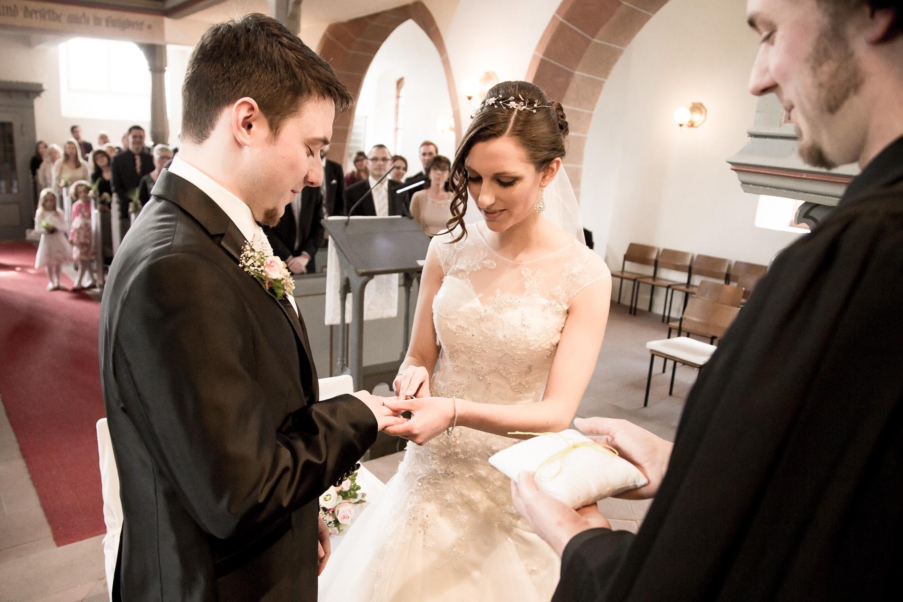 Die Braut steckt den Ehering auf den Finger des Bräutigams auf. Alle anwesenden im Kirchensaal stehen und richten ihre Blicke auf den Ring.