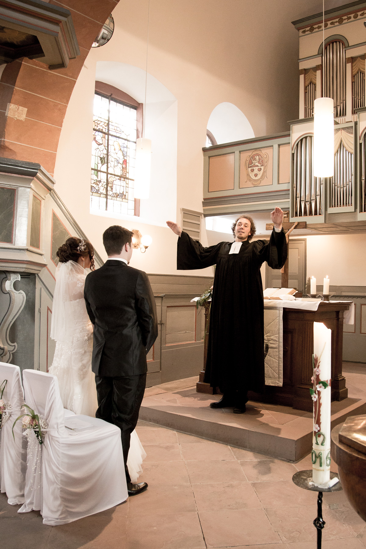 Der Priester streckt seine Arme diagonal hoch, und spricht den Glückssegen dem jungen Hochzeitspaar aus, während sie vor ihm stehen.