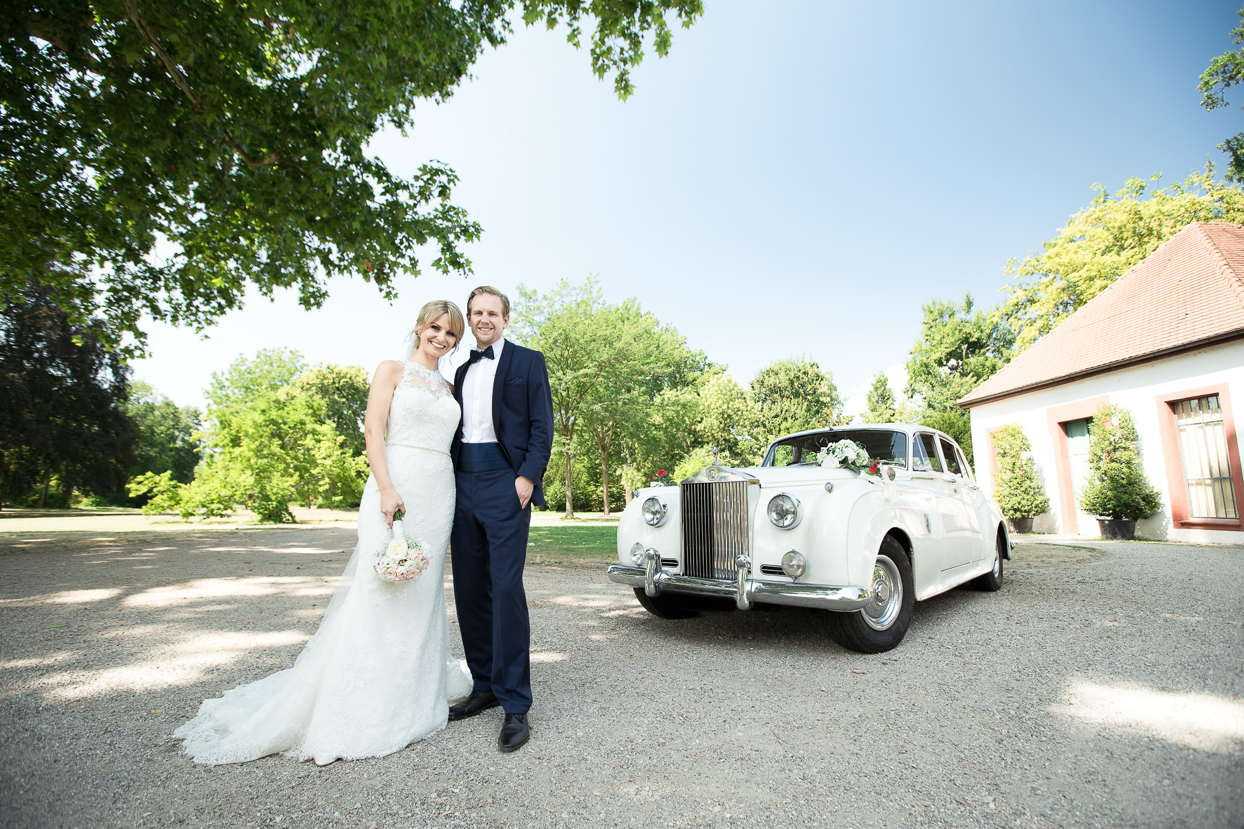 Ein Brautpaar posiert in klassischer Paarpose vor einem Rolls-Royce Oldtimer im Garten des Schlosses Neckarhausen.