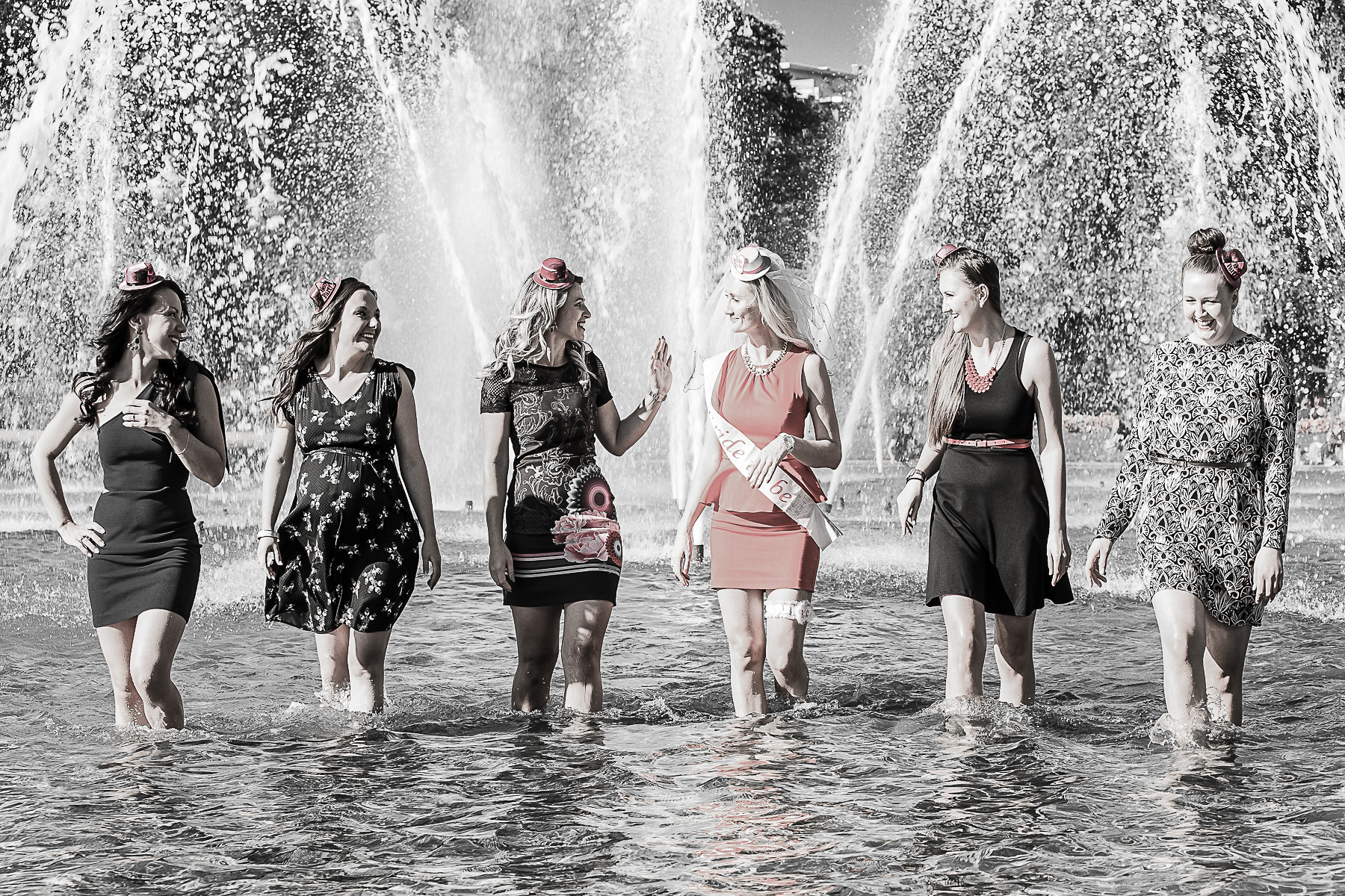Ein Outdoorgruppenbild von Mädels während eines Jungesellenabschieds. Die Junggeselebnbraut und ihre Mädels laufen knietief im Wasserbecken auf die Kamera zu, während sie sich gegenseitig anschauen. Im Hintegrund sind Fontänen sichtbar.