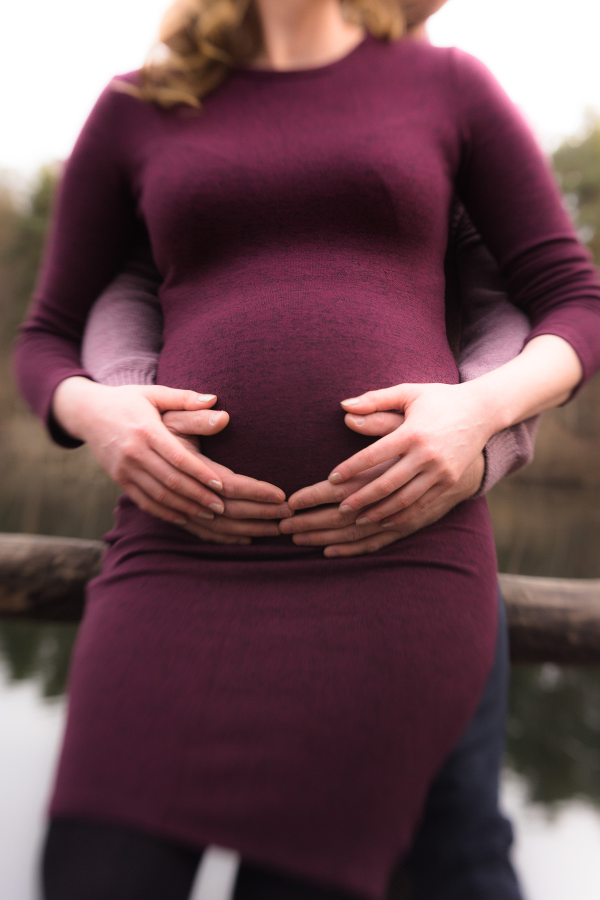 Der Babybauch einer schwangeren Frau im dunkel purpurrotem Kleid frontal, während der Bauch von hinten durch die Hände eines Mannes umfasst wird. Die Frau hat ihre Hände auf die Hände des Mannes aufgelegt.