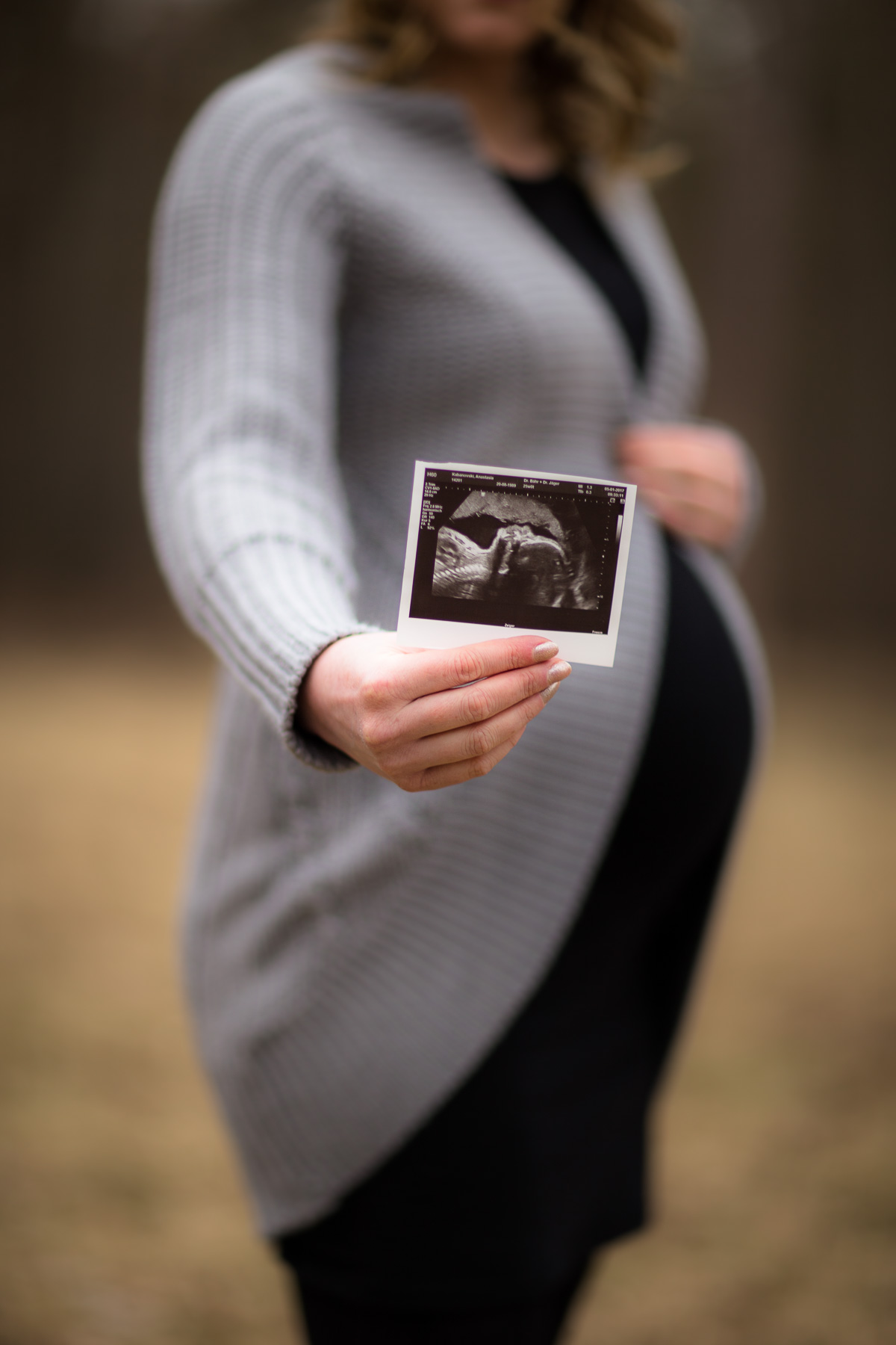 Babybauch-Fotoshooting: Eine schwangere Frau hält mit Ihrer rechten Hand einen Unltraschallabzug ihres noch ungeborenen Babys fest. Im Hintergrund ist Ihr Babybauch und Oberkörper bis zum Kinn sichtbar.