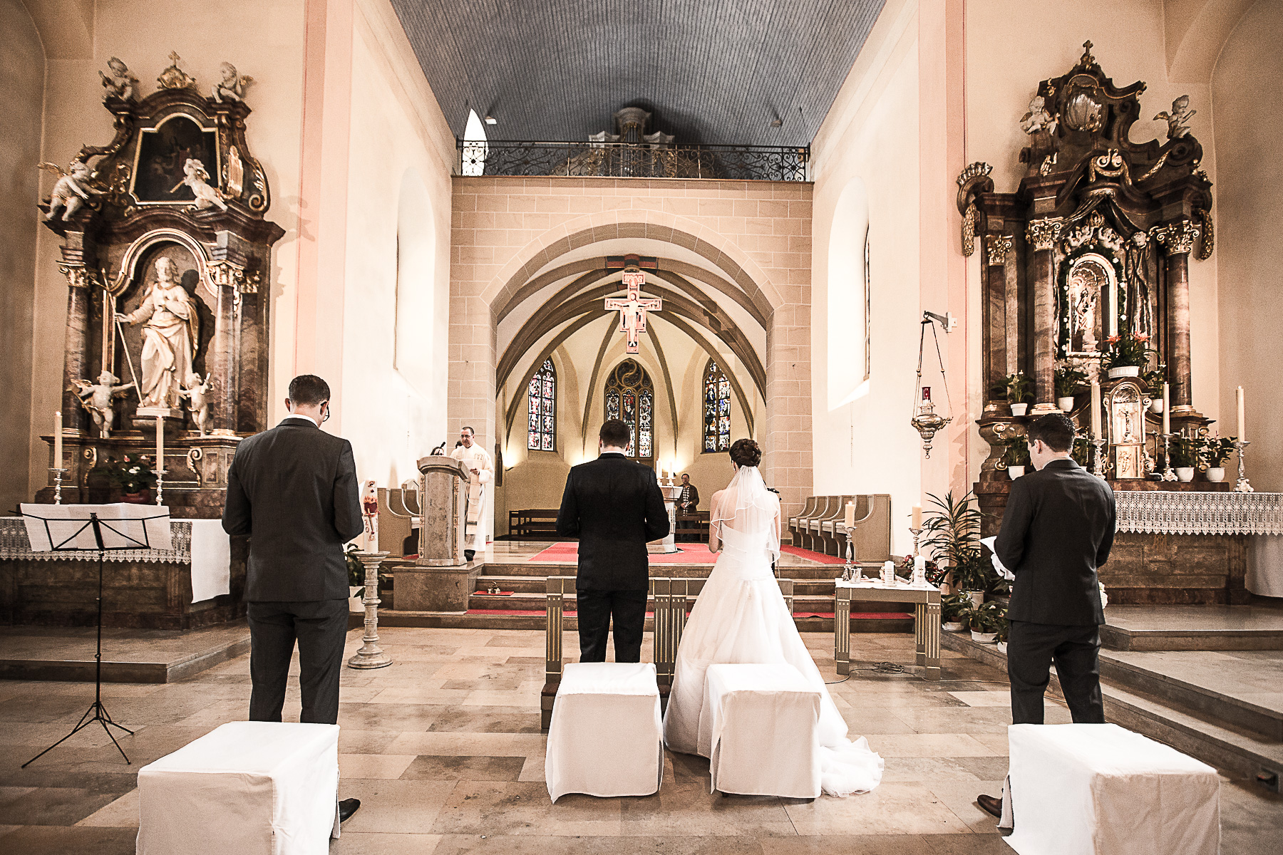 Auf beiden Seiten neben dem Brautpaar stehen die Trauzeugen und lesen die Kirchenliedtexte auf dem Blatt, den Sie in ihren Händen halten. Im Hintergrund ist der Priester vor seinem Lesepult auf dem Ambo sichtbar.
