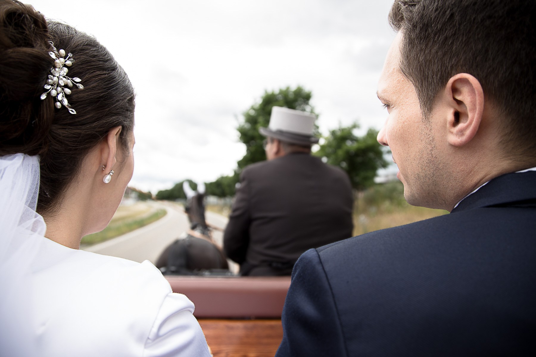 Das Brautpaatpaar sitzt auf der Kutsche Schulter an Schulter. Im Hintergrund fährt der Kutscher seine Pferde.