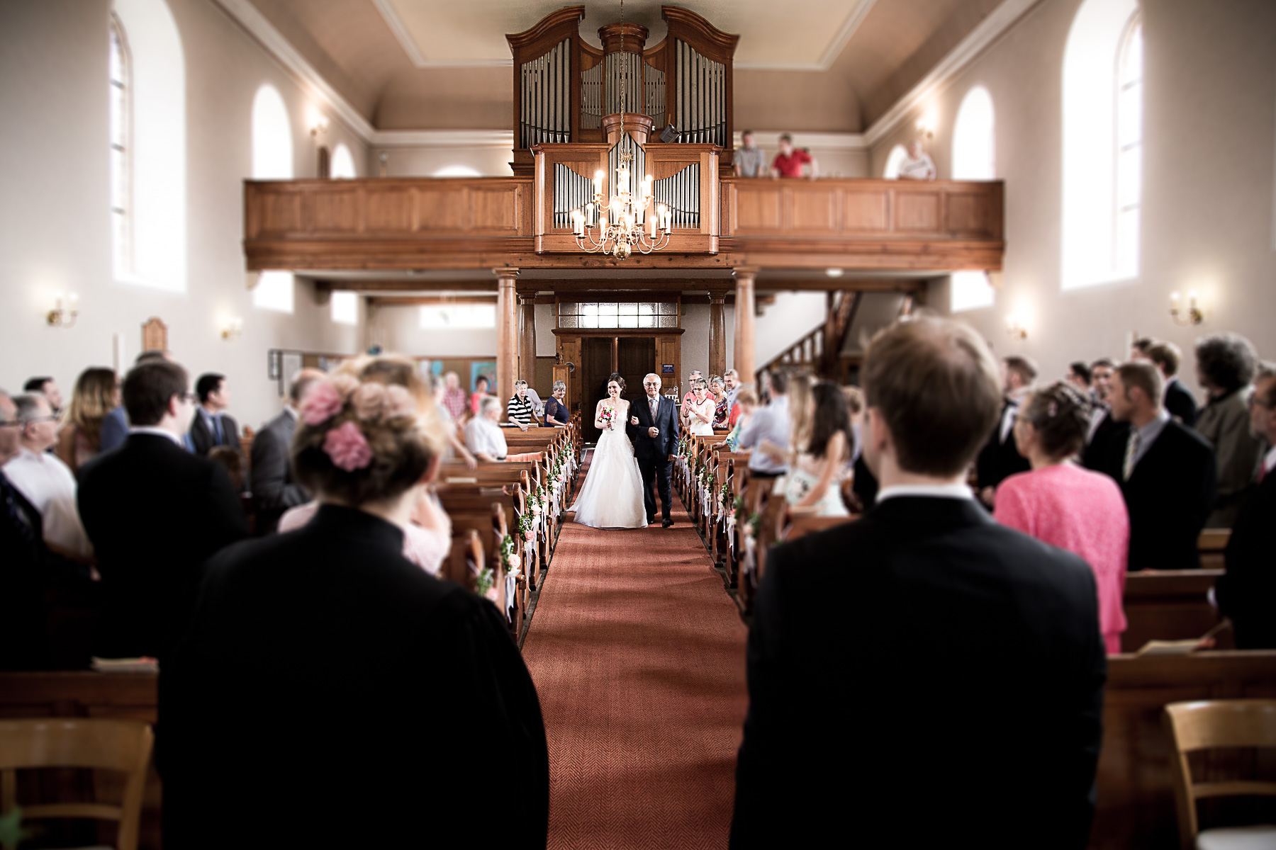 Die Braut hält sich an ihrem Vater fest und lächelt, während sie den Hauptgang des Kirchensaals passieren. Sie sind bereits auf dem halben Weg zum Bräutigam. Der Bräutigam, die Priesterin und alle Traugäste haben ihre Blicke auf die Braut gerichtet.