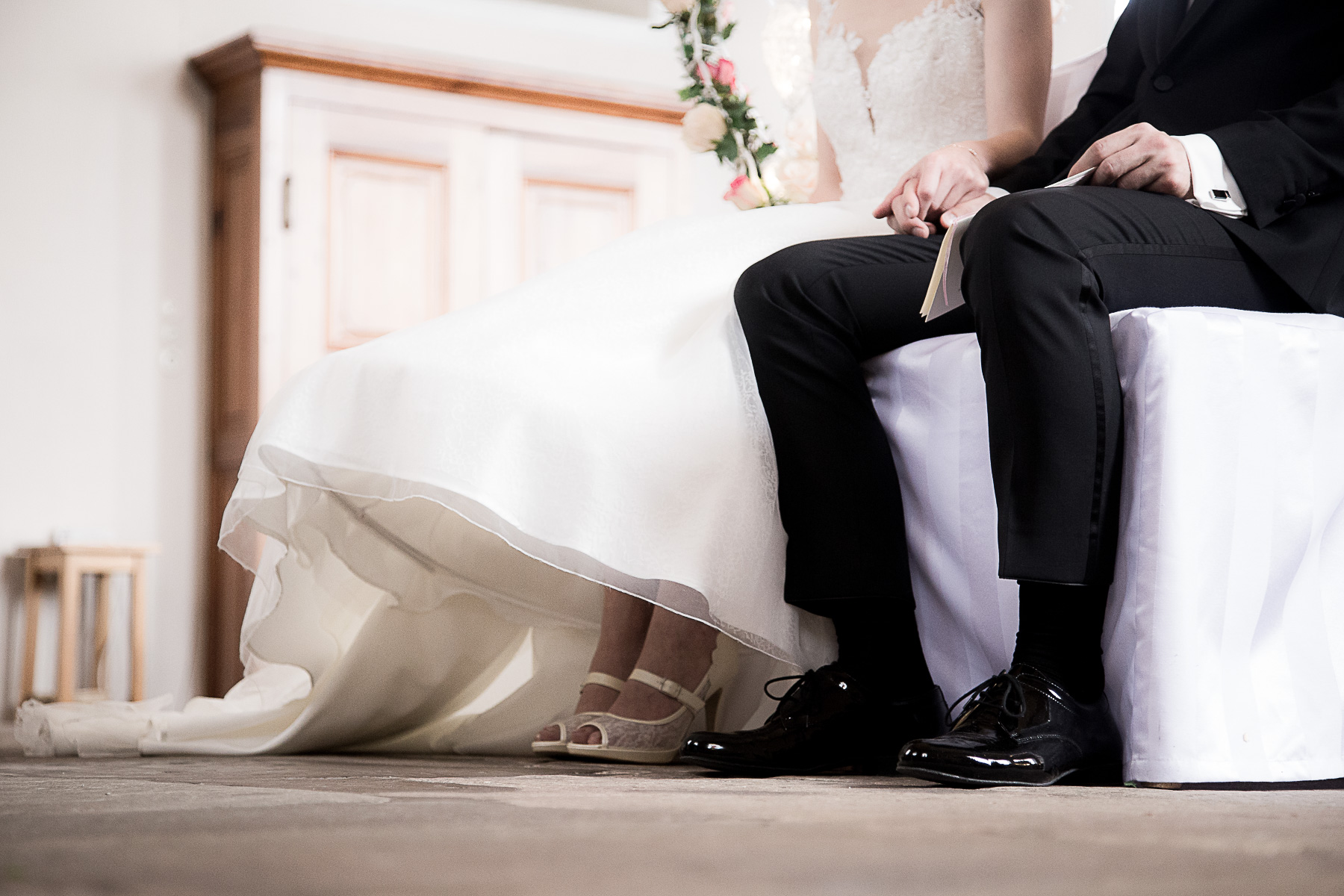 Der untere Teil Hochzeitspaars inklusive ihrer Schuhe. Sie sitzen auf den Traustühlen nebeneinander und halten Händchen.