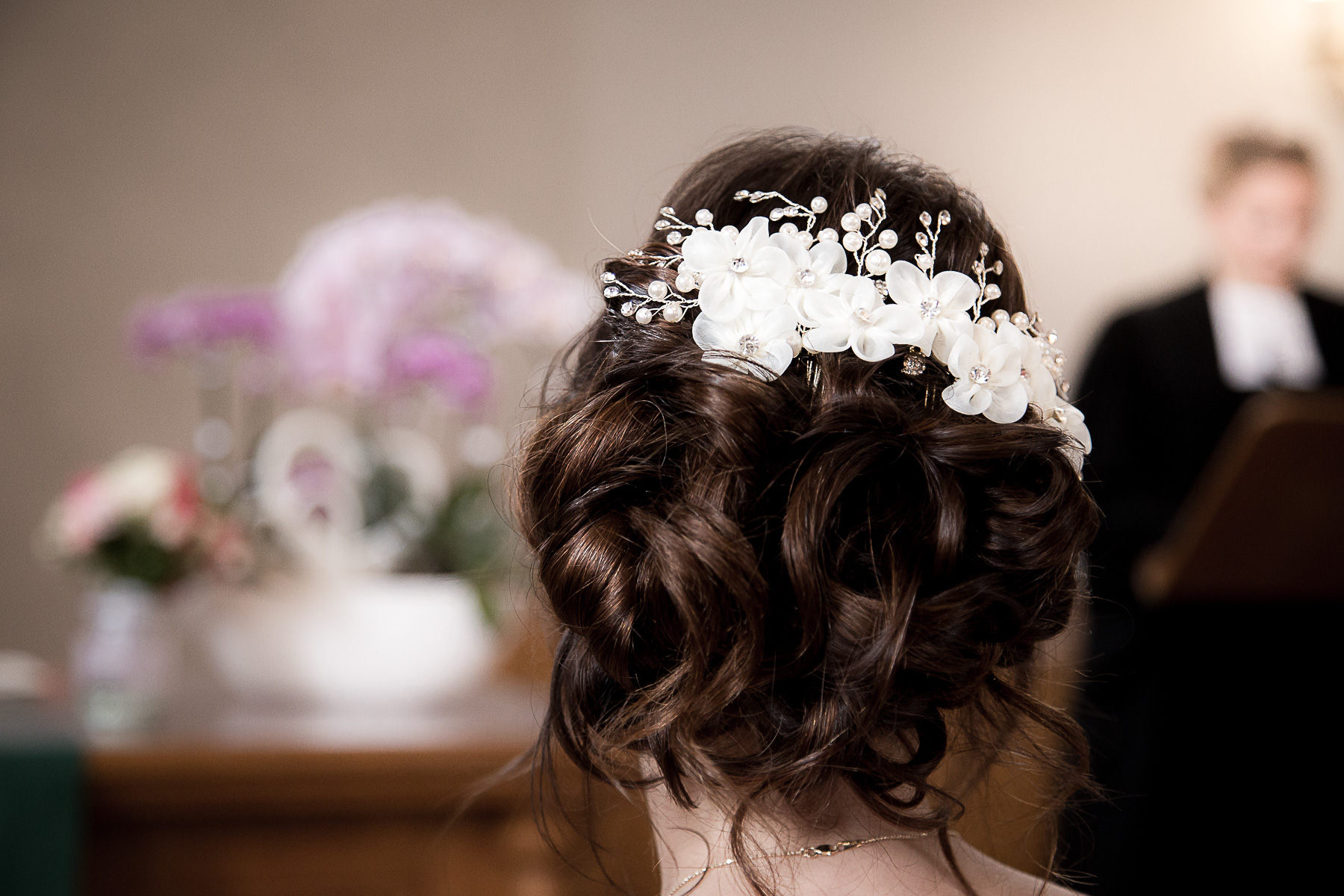 Eine Detailaufnahme der Frisur von der Braut aus der Rückansicht. Sie hat eine elegante Chignon-Frisur mit einer weißen Haarbrosche.