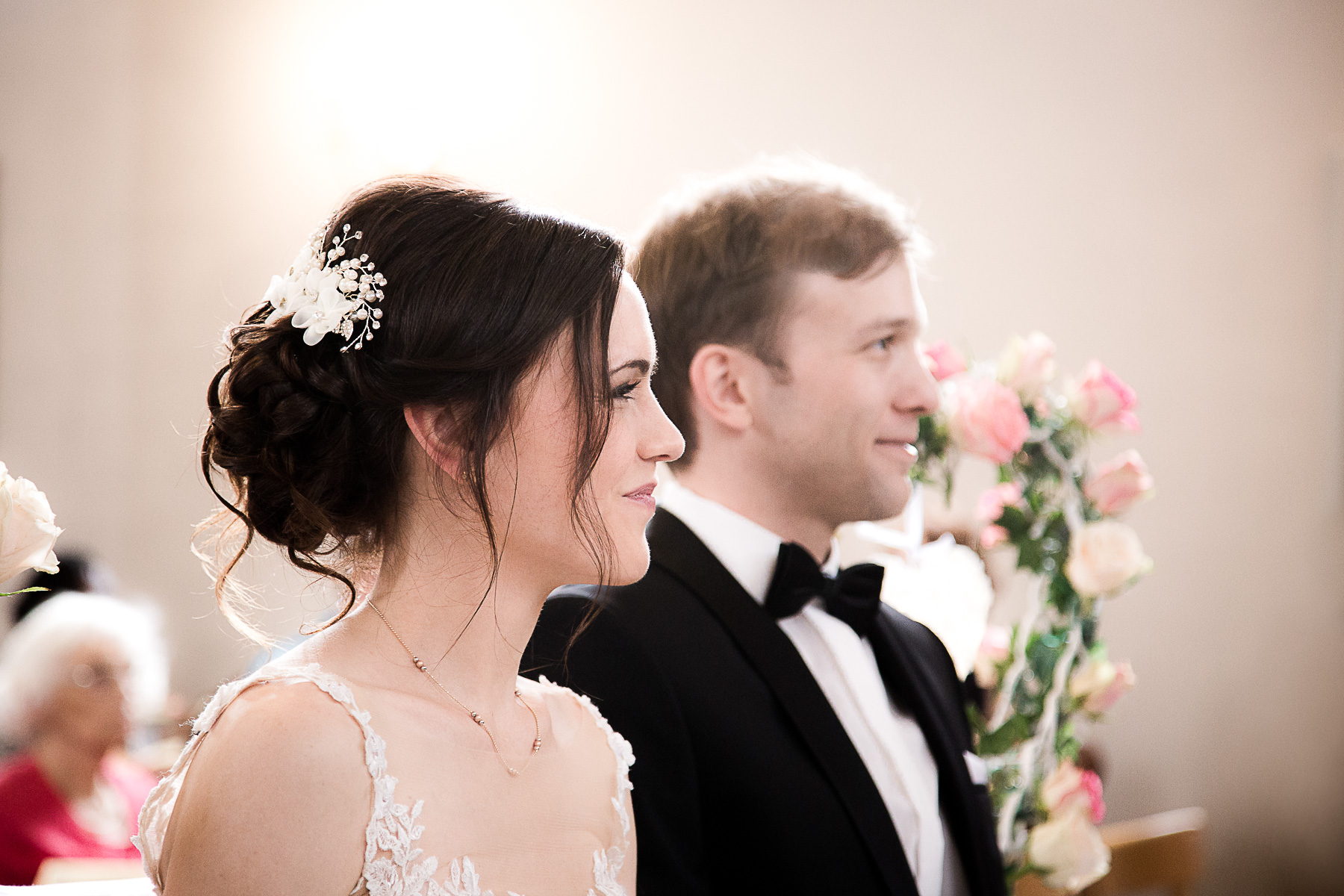 Die Profilnahansicht der Braut, während sie lächelt. Direkt hinter ihr ist des Profil des Bräutigams leicht verschwommen sichtbar. Er lächelt ebenfalls.
