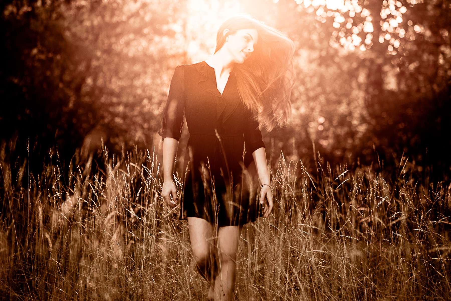 Eine junge Frau läuft dynamisch im hohen Rasen und wirft ihre langen Haare mit dem Kopf zur Seite. Sie wird von hinten mit den warmen Strahlen der Abendsonne umhüllt.