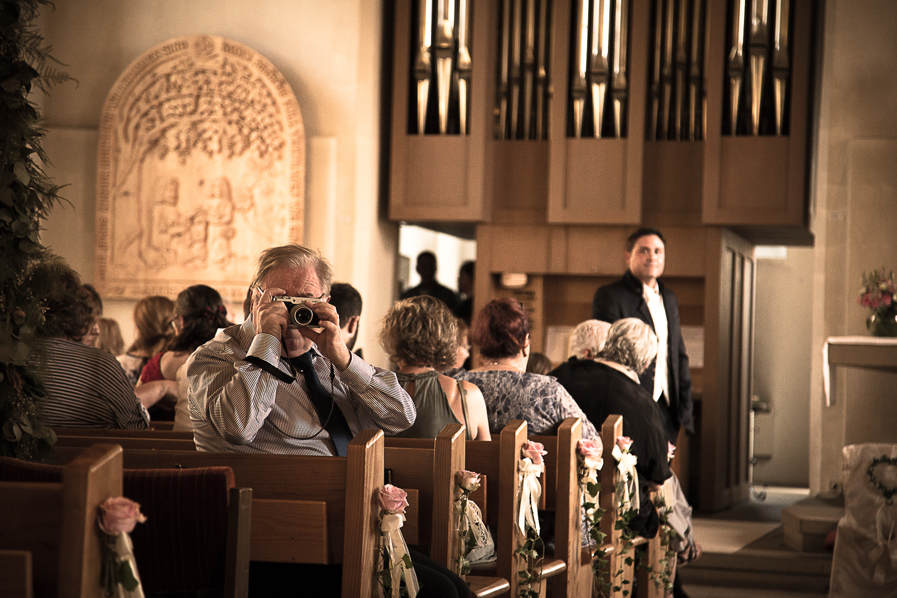 Ein Gast hat sich mit seiner Kamera in Stellung gebracht, um die Braut beim Eintritt in den Kirchensaal zu fotografieren, während im Hintergrund der Bräutigam vor dem Altar warten.