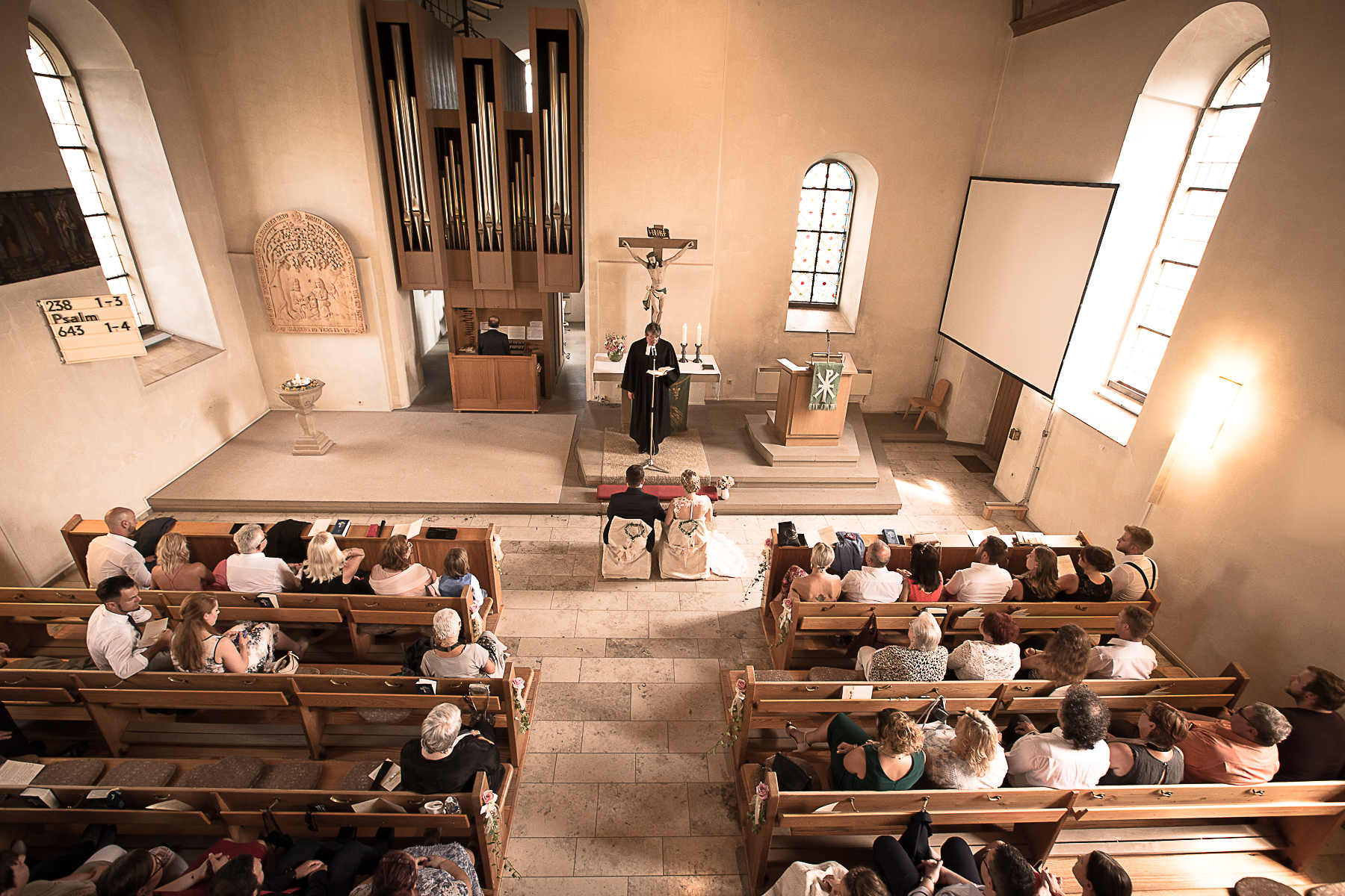 Die Vogelperspektive einer kirchlichen Trauung von der Orgelempore. Der Priester steht vor dem Altar und spricht Richtung Brautpaar. Alle Gäste hören hören zu, während sie auf den Kirchbänken sitzen.