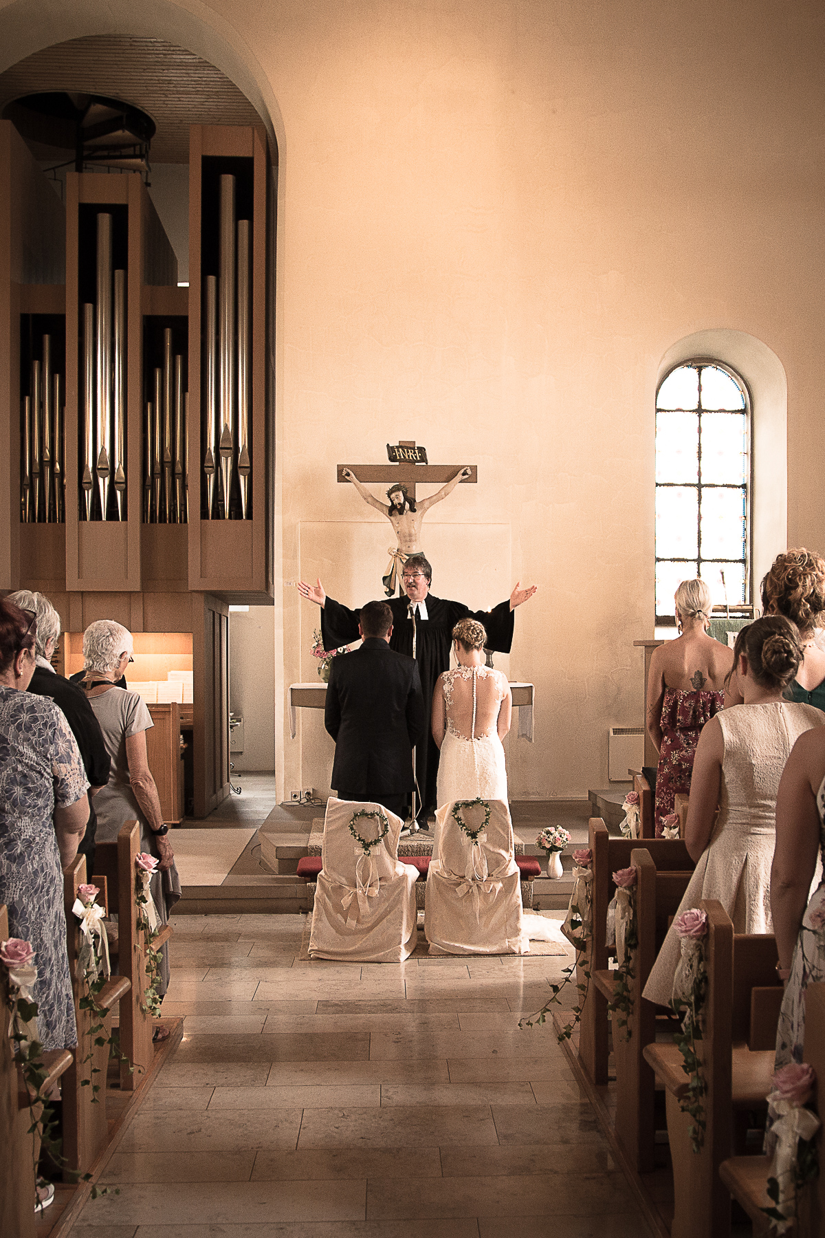 Während einer kirchlichen Trauung traut der Priester das Brautpaar, in dem er seine Hände über ihre Köpfe hochhält. Währenddessen stehen alle aufrecht, auch die Gäste.