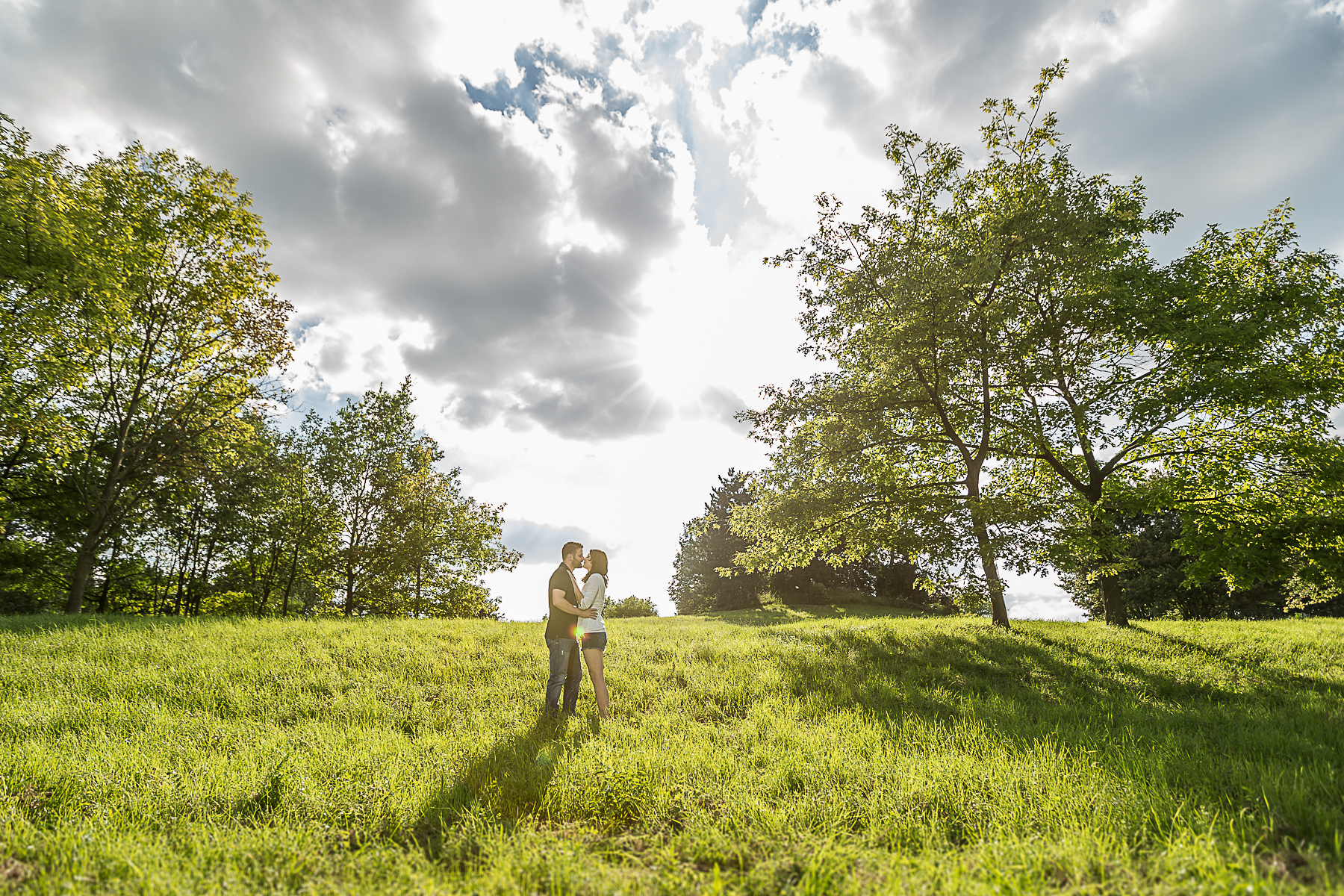 Ein junges Paar steht in einem hügligen Feld und küsst sich. Beidseitig sind Bäume, im Himmel sehr bewölkt und die Sonne scheint durch die Wolken hindurch.