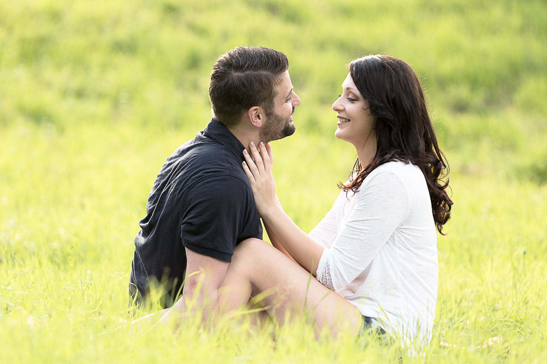 Ein Liebespaar sitzt in einem hellgrünen Feld nah bei einander. Sie lächeln und schauen sich in die Augen. Die Frau fasst den Mann an seinem Hals an.