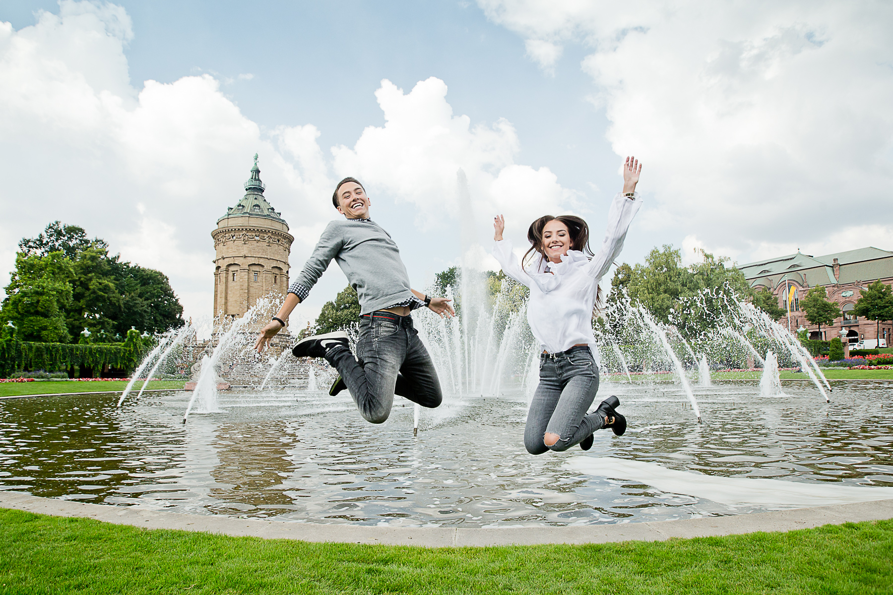 Ein junges Paar springt energievoll hoch, sodass ihre Beine angewinkelt sind. Sie lachen. Im Hintergrund sind Fontänen und der Wasserturm von Mannheim.
