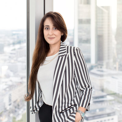 Busness-Foto: Eine in Business Casual gekleideten junge Frau. Im Hintergrund, hinter dem Fenster, sind die Hochhäuser des Bankenviertels von Frankfurt.