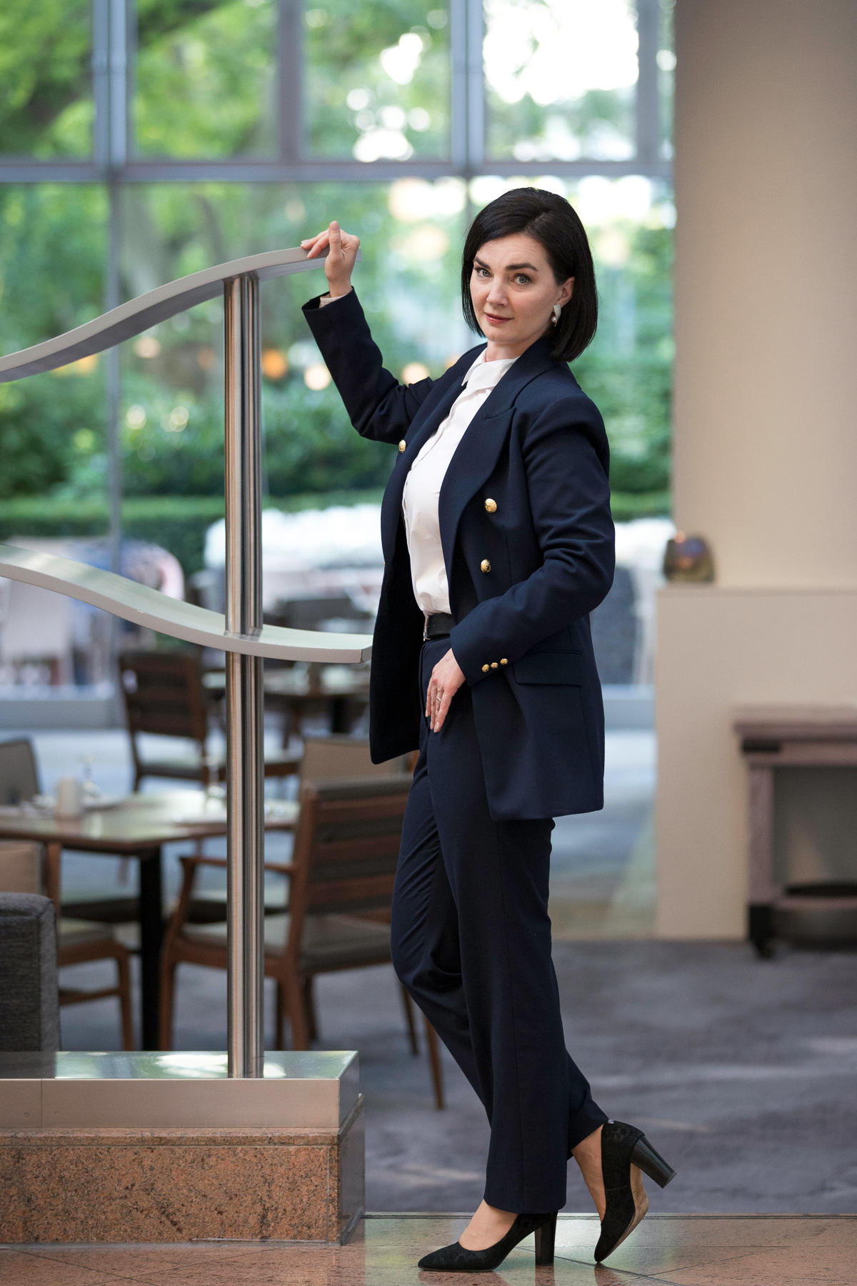 Eine klassisch-businessgekleidete Frau in Ganzkörperaufnahme. Sie hält sich an einer Edelstahlbrüstung mit einer Hand fest und steht in einer lockeren Pose. Im Hintergrund das Restaurant eines Hotels sowie grüner Garten hinter den Fenstern.