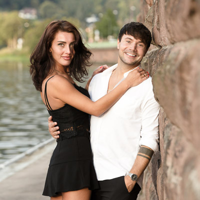 Ein junges Paar posiert neben einem Fluss und einer Sandsteinmauer. Sie halten sich gegenseitig fest und lächeln in die Kamera.